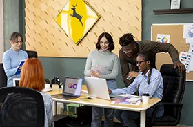 3 people looking at a laptop screen and 2 other woman working inside an ad agency office