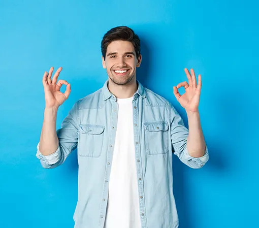 Confident smiling young man in denim jacket showing ok signs with both hands standing against blue background