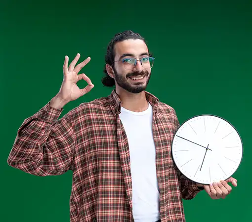 Smiling young handsome cleaning guy wearing t-shirt holding wall clock showing okay gesture isolated green wall