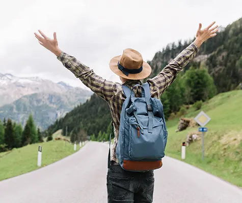 Joyful male traveler standing on a highway with his hands raised, looking towards distant trees.